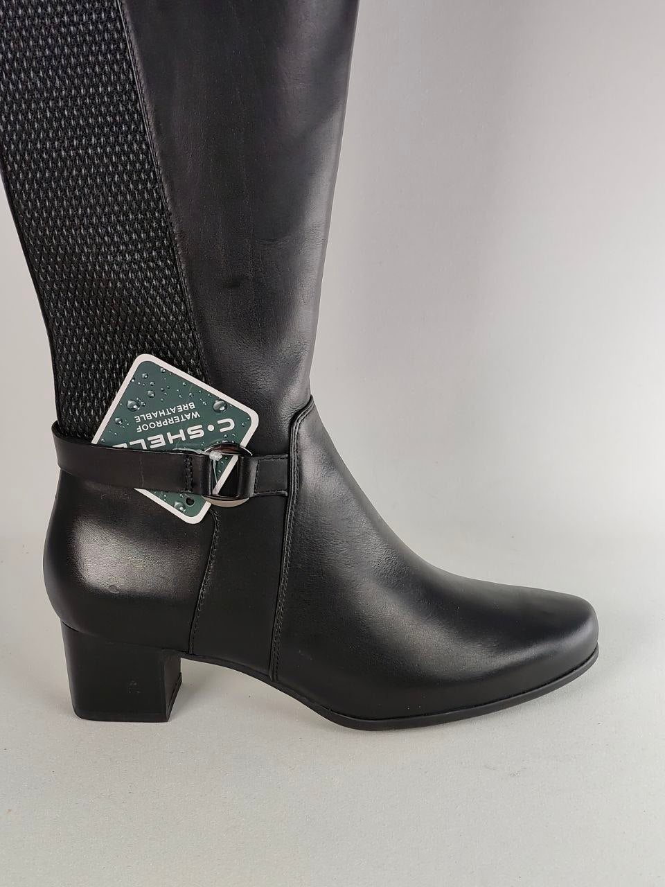 A black knee-high boot with a smooth leather-like finish, a textured black mesh panel on the upper side, and a buckle strap detail around the ankle. A green and white C.