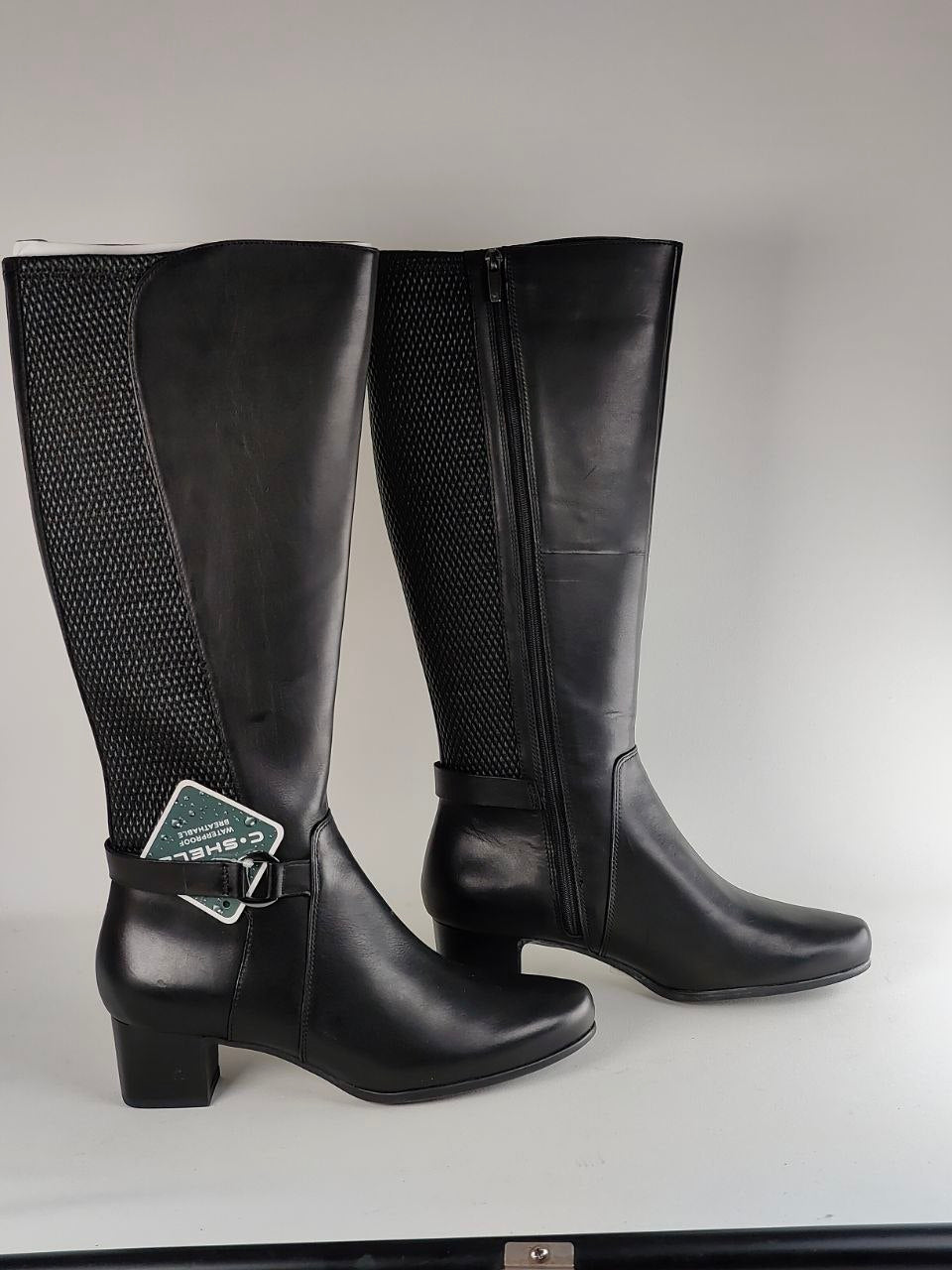 A pair of black knee-high riding boots with a smooth leather shaft, perforated side panels, and a low block heel, shown against a neutral gray background.
