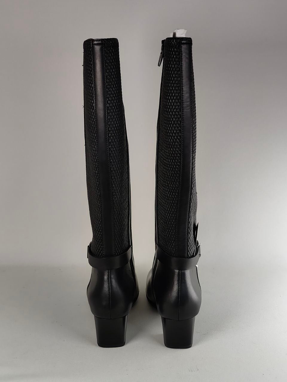 A pair of black knee-high boots shown from the back, featuring a textured pattern on the shaft and a smooth black leather lower section with block heels, set against a neutral gray background.