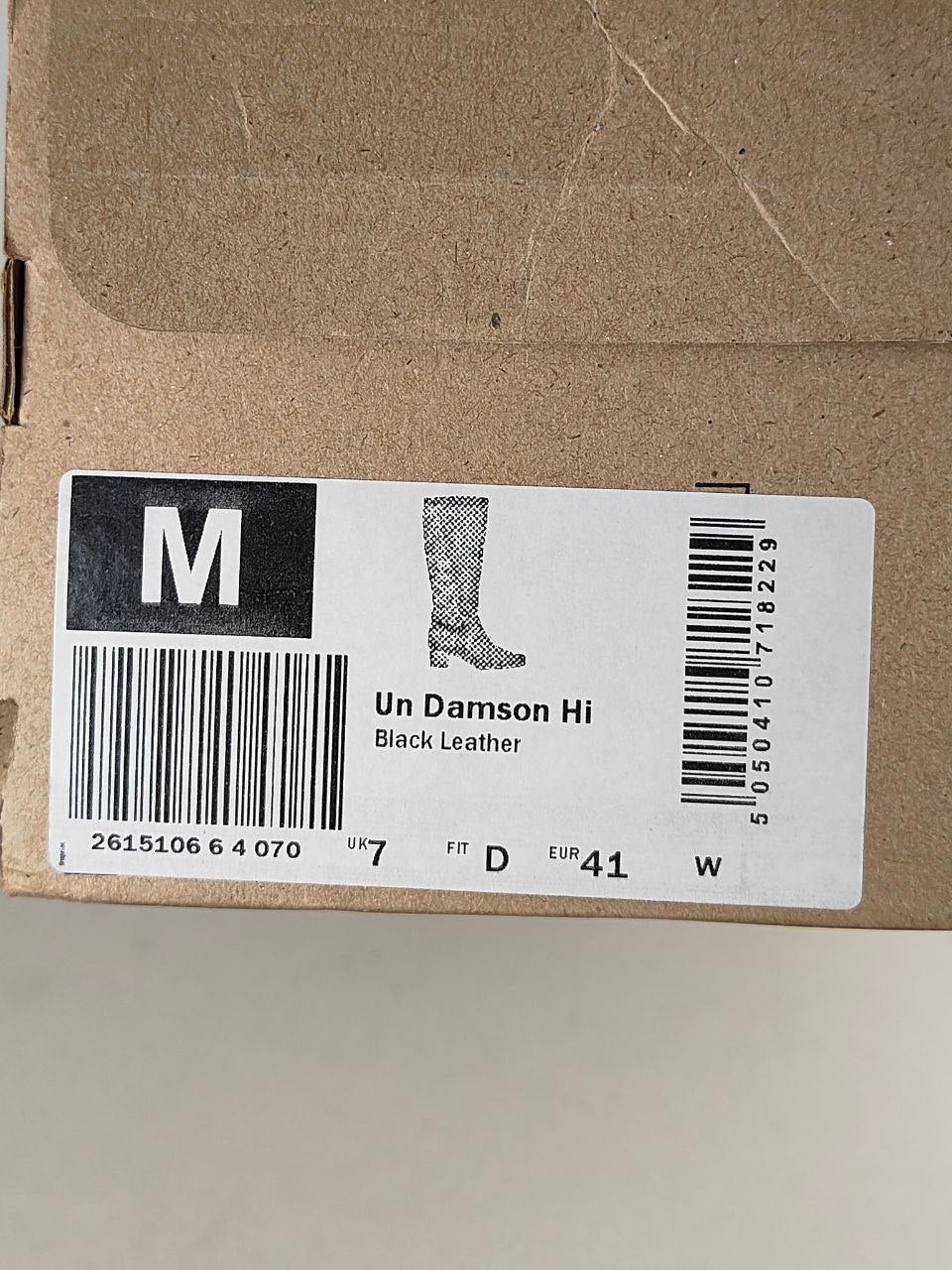 A close-up of a brown cardboard shoe box with a white size label. The label shows a black block with a white letter M, a barcode, a simple line drawing of a knee-high boot, and the text 'Un Damson Hi' and 'Black Leather'.