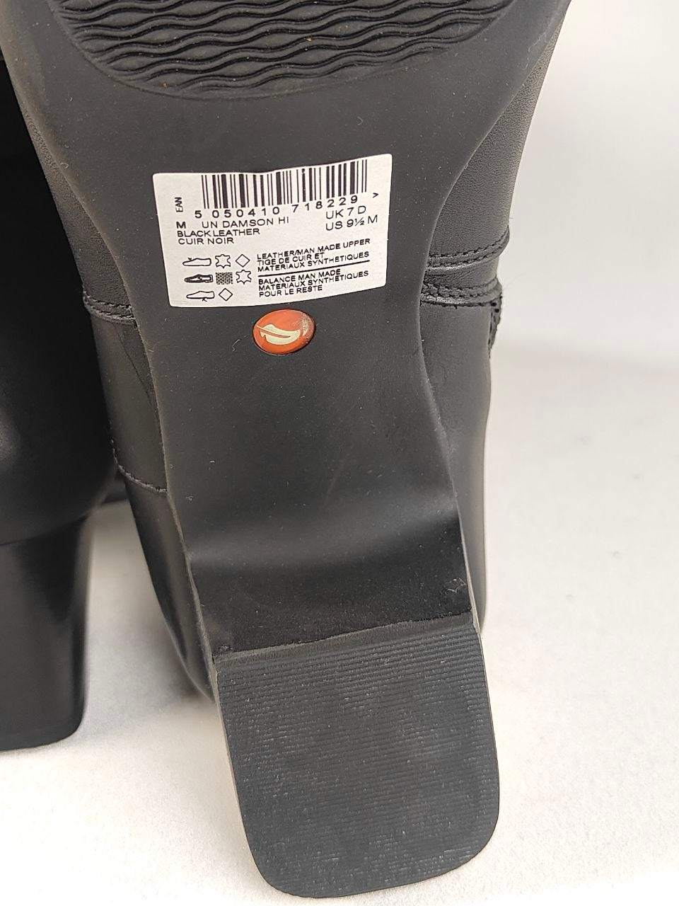 Close-up of the sole of a black boot, showing a white size label with barcode and a small orange circular logo, photographed on a light gray background.
