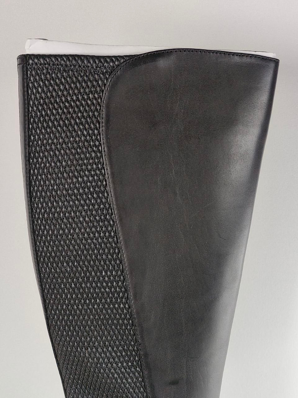 A close-up of a black knee-high boot featuring a smooth leather side panel and a textured woven pattern panel on the other side, set against a light gray background.
