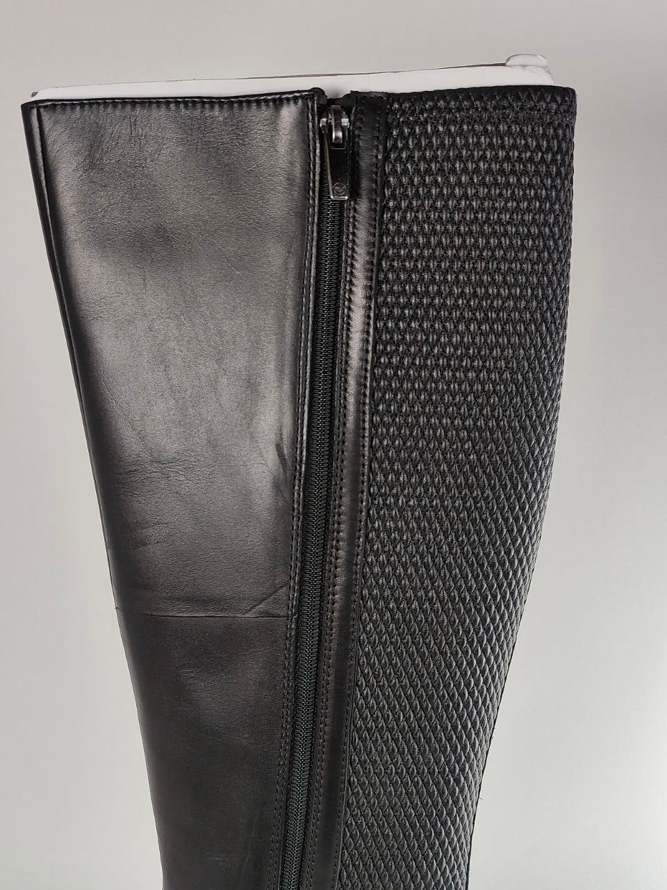 Close-up of a black knee-high boot showing a side zipper, a smooth leather-like panel on the left, and a textured woven pattern on the right.