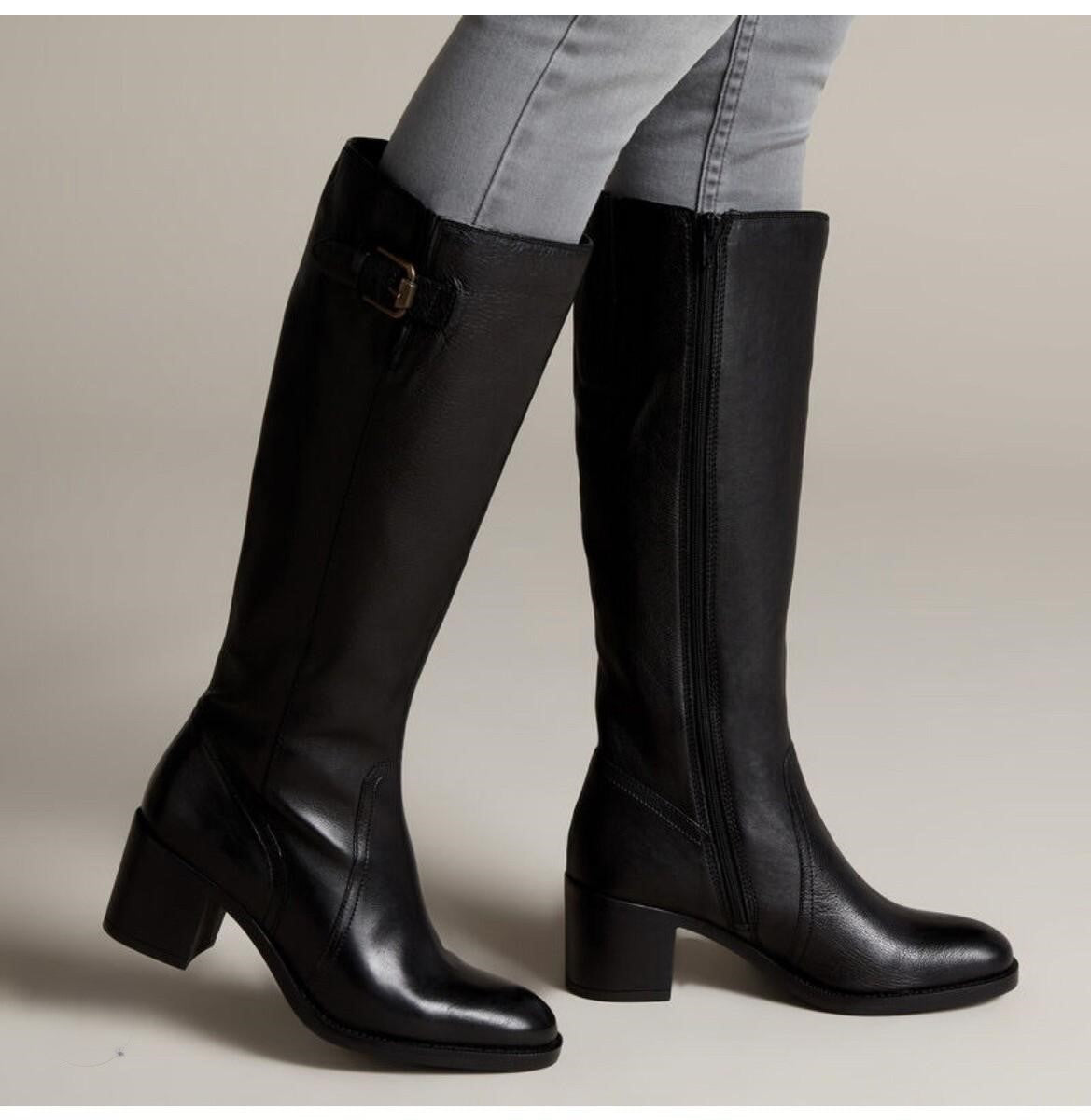 A pair of black leather knee-high boots with block heels, shown on a model wearing light gray jeans against a neutral beige studio background.