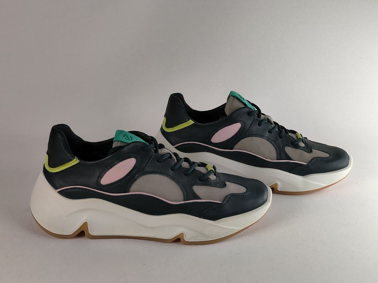 A pair of chunky low-top sneakers in black, gray, and white, featuring light pink accents and lime green details, photographed on a neutral gray background.
