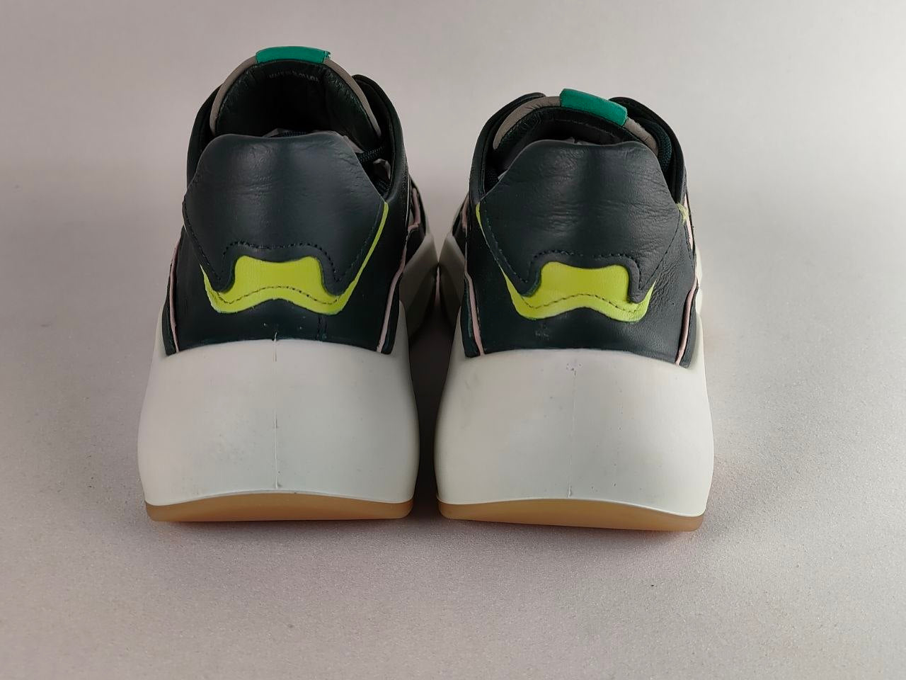 A pair of low-top sneakers shown from the back, featuring dark gray leather uppers with a bright yellow-green accent, thick white midsoles, and gum soles.