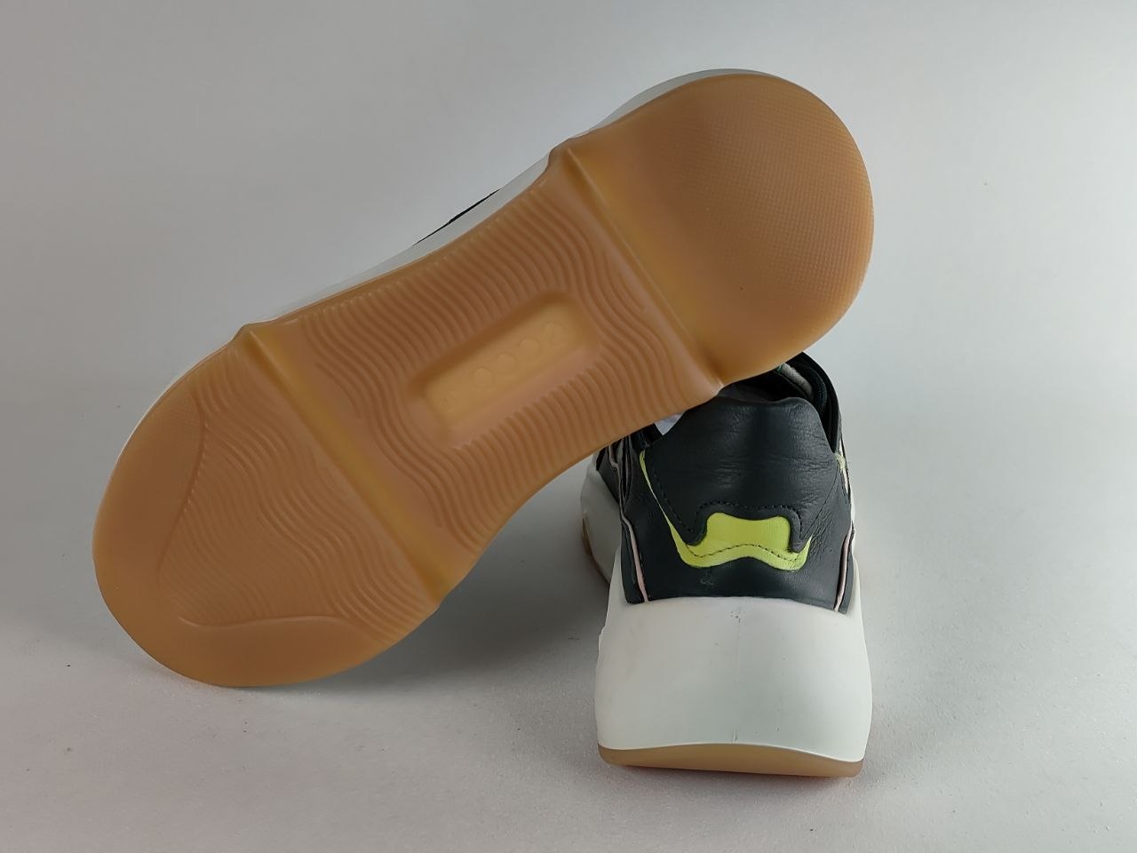 A black and white sneaker with a bright yellow-green accent on the heel, shown from the back with its tan outsole propped up against a light gray background.