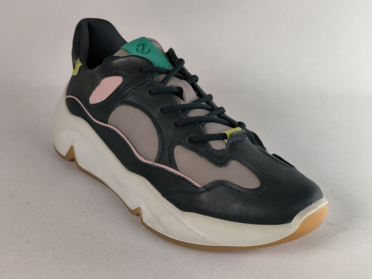 A low-top sneaker with a black leather upper, light gray mesh panels, a thick white midsole, and pastel pink accents, photographed on a neutral gray background.