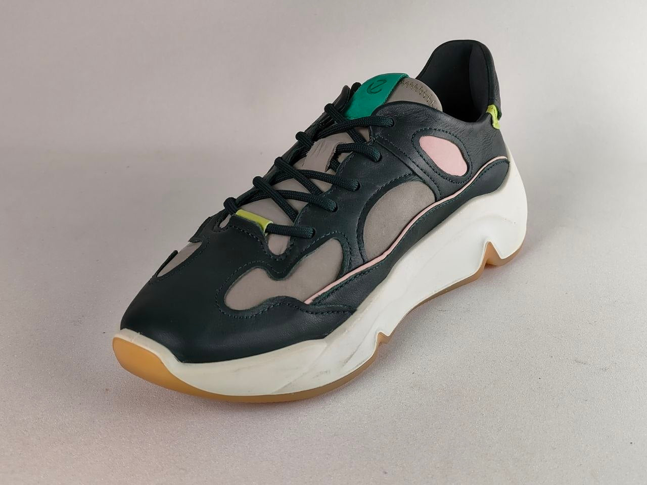 A single low-top sneaker shown at a three-quarter angle on a neutral gray background. The shoe features a black leather upper with gray mesh panels, a thick white chunky sole, a light pink side accent, and a gum rubber outsole.