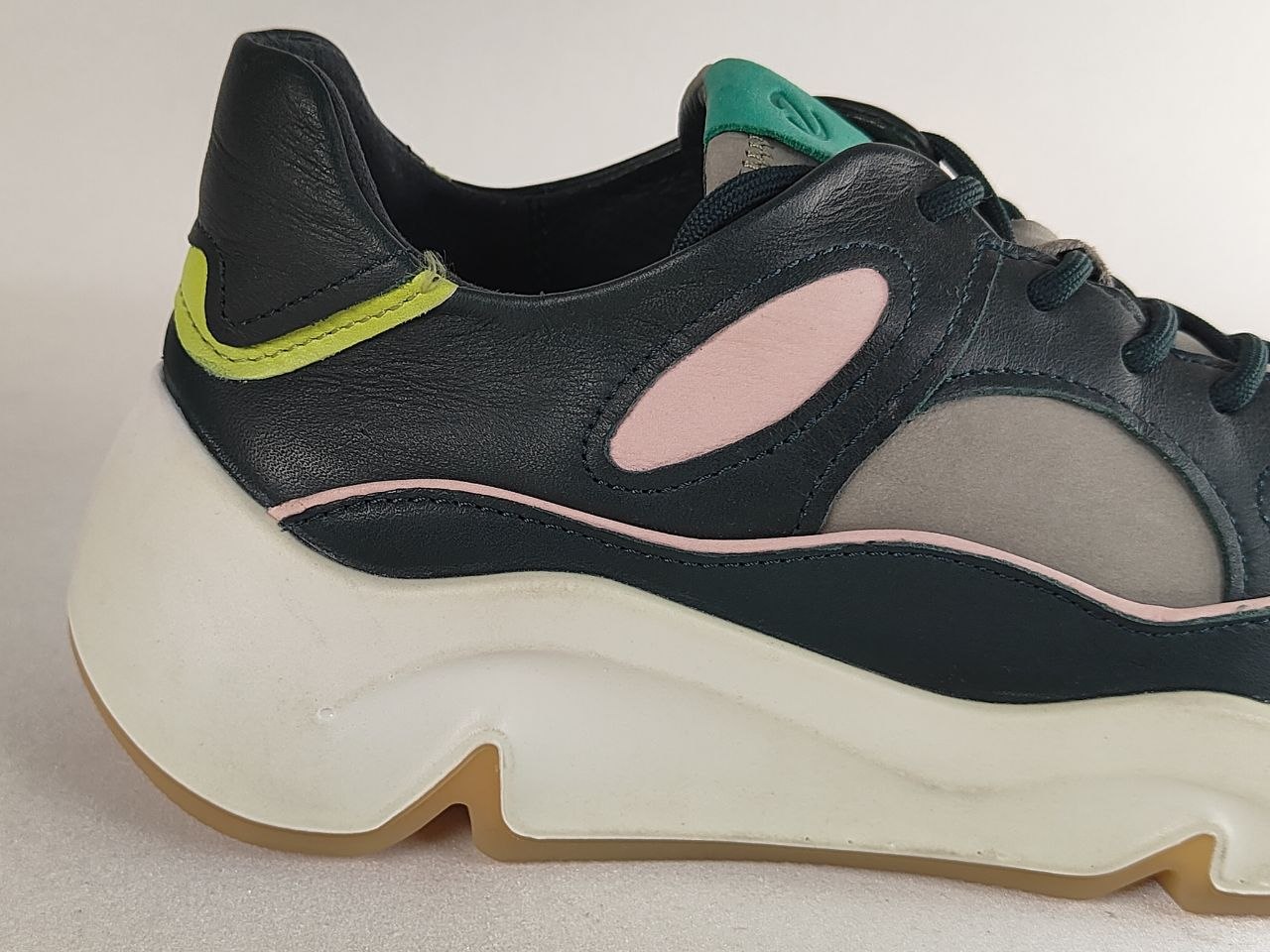 A close-up side view of a chunky platform sneaker featuring a black leather upper with light pink and gray mesh panels, a lime green heel accent, a white sculpted sole, and a gum outsole.