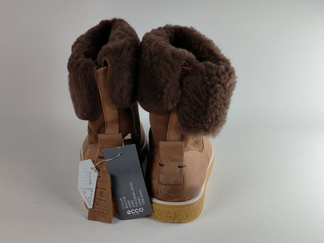 A pair of ECCO mid-calf boots in tan leather with dark brown shearling-style fleece trim, shown from the back with product tags attached, on a light gray background.