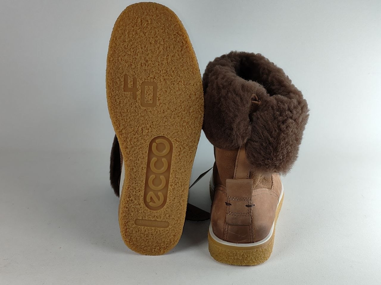 A pair of brown leather and shearling mid-calf boots, showing the textured tan sole of one boot and the side profile of the other with a fluffy brown shearling collar.