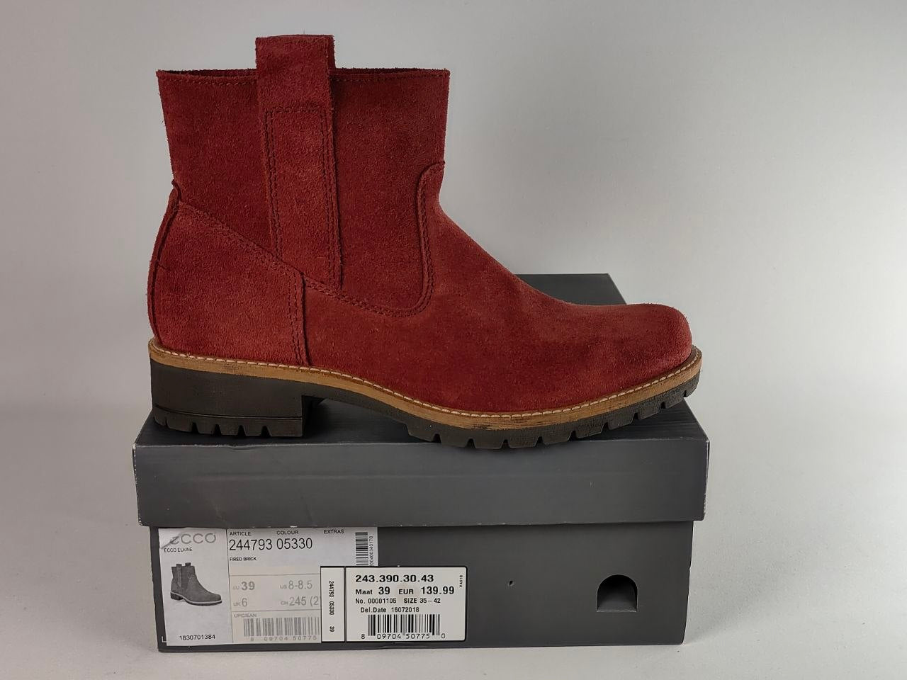 A red suede Chelsea-style ankle boot with a brown sole, displayed on top of a dark gray shoebox against a light gray background.