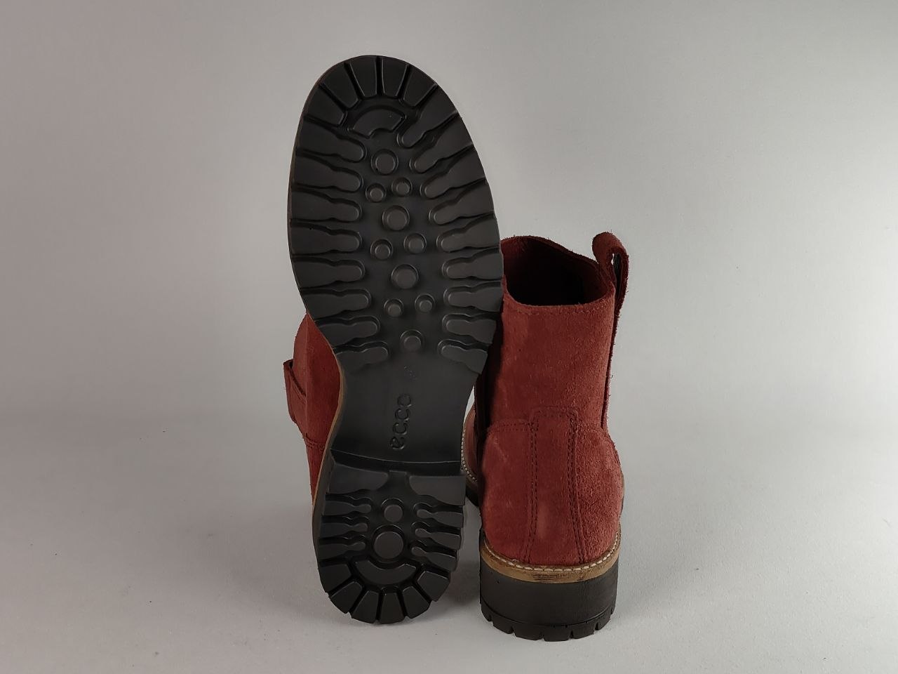 A pair of red suede ankle boots shown from the back, with one boot standing upright to display the red suede shaft and the other lying flat to show the black treaded outsole.