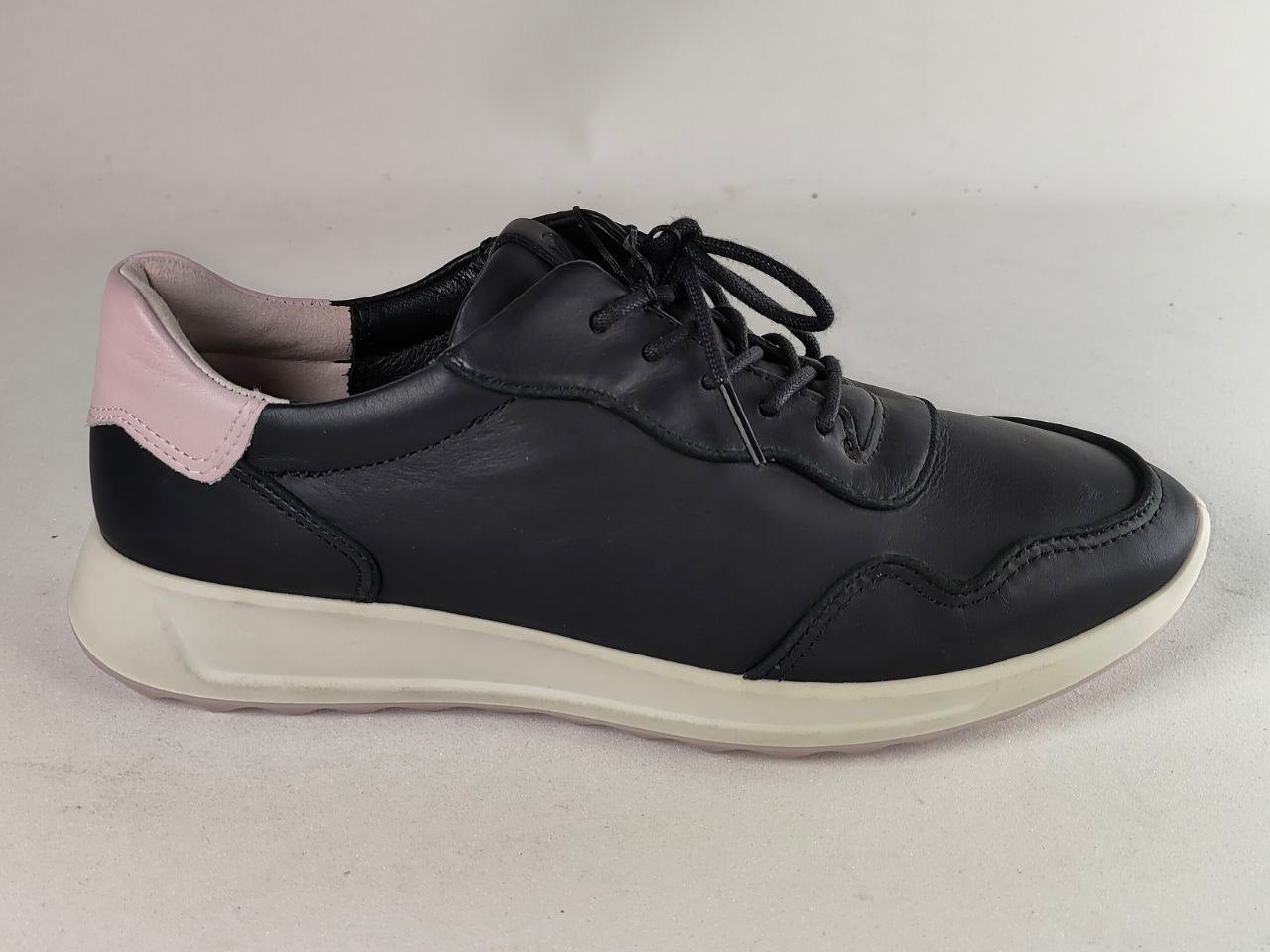 A black leather low-top sneaker with a thick off-white sole and a light pink heel tab, shown from a side profile on a neutral light gray background.