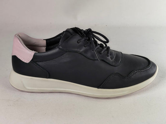 A black leather low-top sneaker with a thick off-white sole and a light pink heel tab, shown from a side profile on a neutral light gray background.