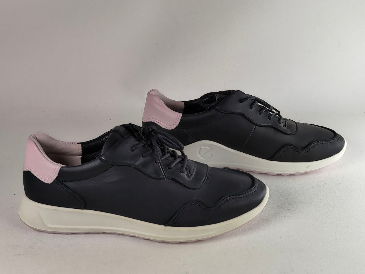 A pair of black leather low-top sneakers with light pink heel accents and white soles, photographed on a neutral light gray background.