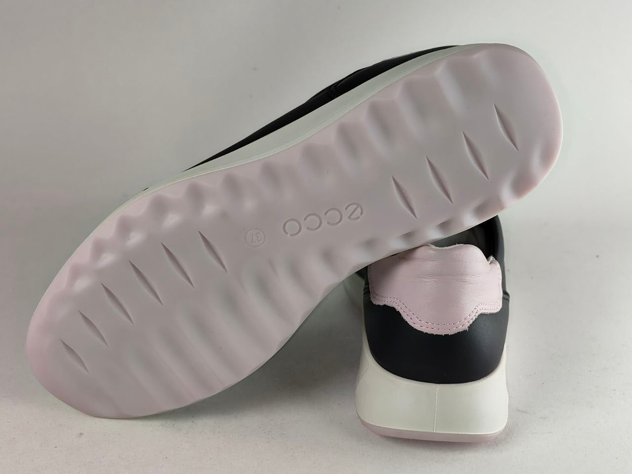 A close-up of an ECCO sneaker showing the light pink textured outsole and the side profile of the shoe which features a black upper, light pink heel tab, and white midsole.