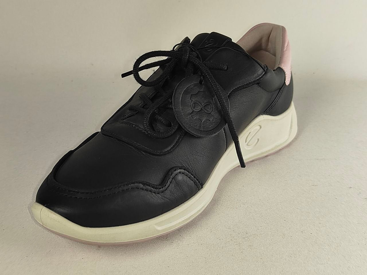 A black leather sneaker with black laces, a circular black logo patch on the side, and a thick cream-colored midsole and outsole, photographed on a neutral light beige background.
