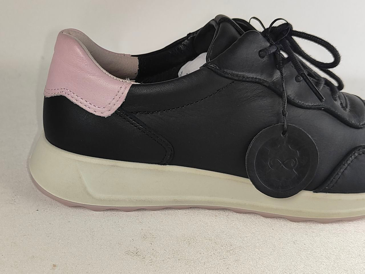 A black leather low-top sneaker with a light pink heel tab and a thick off-white platform sole, shown from a side profile view with a circular hangtag attached.