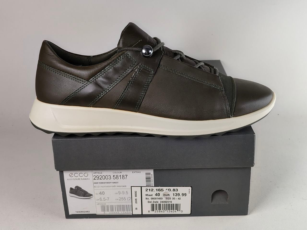 A dark brown leather Ecco low-top sneaker with a white midsole, displayed on top of its dark gray shoebox against a light gray background.