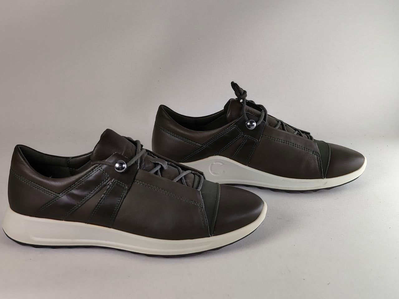 A pair of dark brown leather low-top sneakers with white soles and silver metal eyelets, displayed on a neutral gray background.