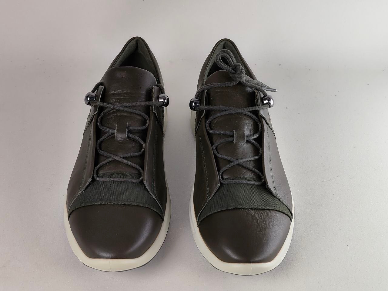 A pair of dark brown leather low-top sneakers with matching dark brown laces, silver metal eyelets, and white soles, shown against a neutral light gray background.