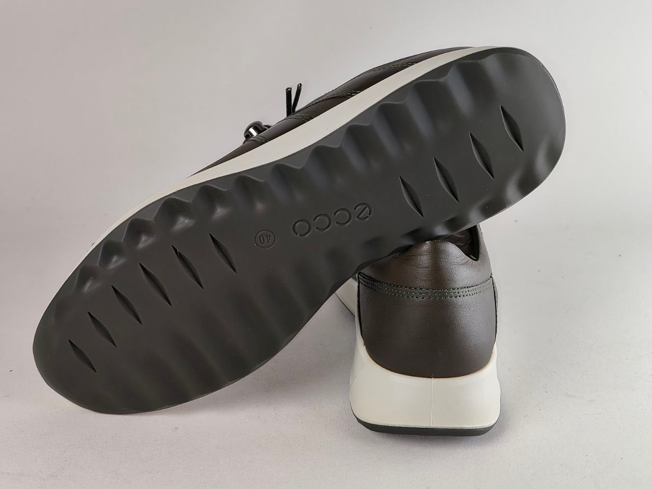 A pair of dark brown leather shoes with a thick white midsole and a black textured outsole. One shoe is positioned upright while the other is tilted on top, showing the 'ecco' brand name embossed on the black sole.
