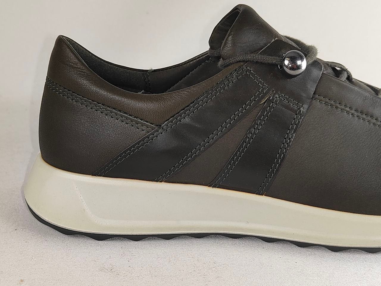 A close-up side view of a dark brown leather sneaker with a thick off-white sole and a silver metal accent on the laces.