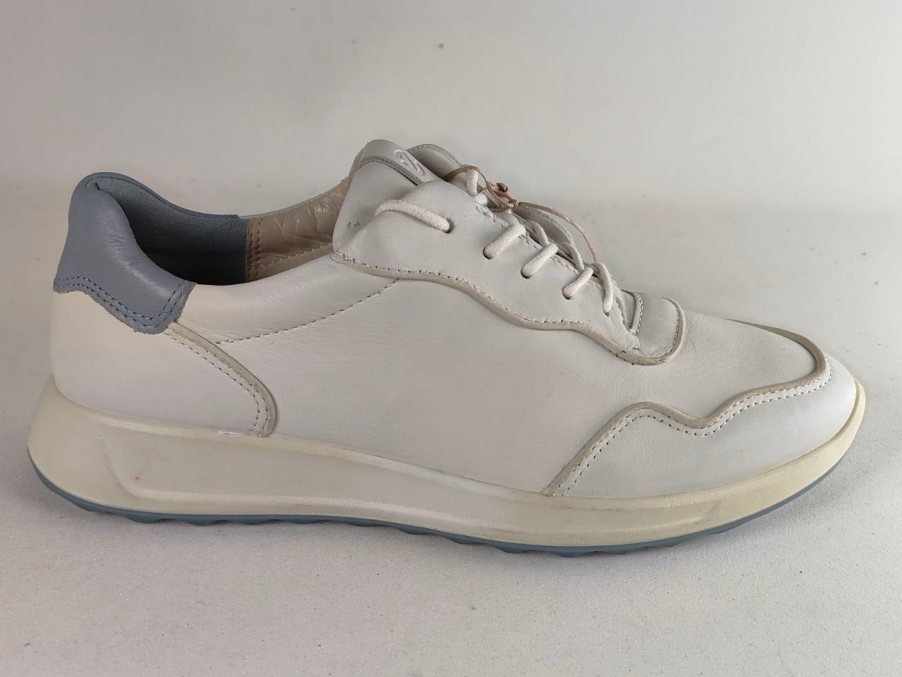 A white leather low-top sneaker with a gray heel tab and a thick cream-colored sole, shown from a side profile view against a neutral light gray background.