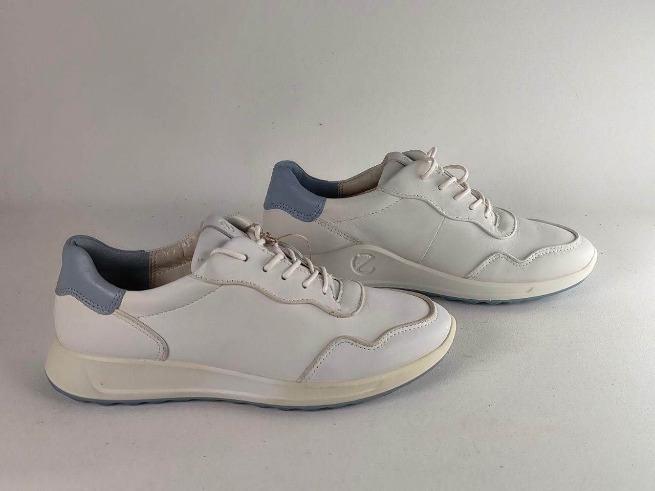A pair of white low-top sneakers with light blue heel accents, shown on a neutral light gray background.