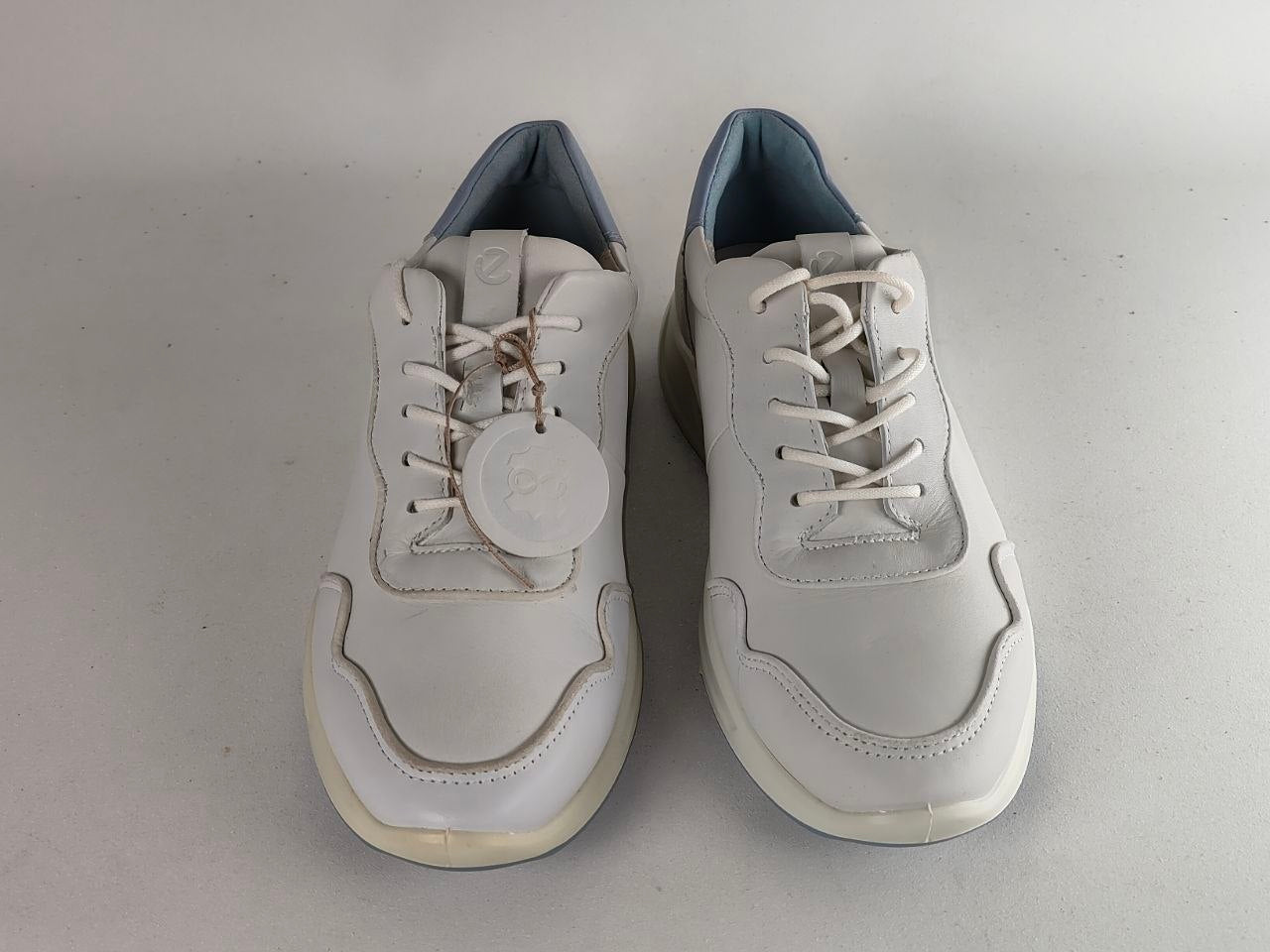 A pair of white leather low-top sneakers with white laces, cream-colored soles, and a light blue-gray lining, shown on a neutral gray background.