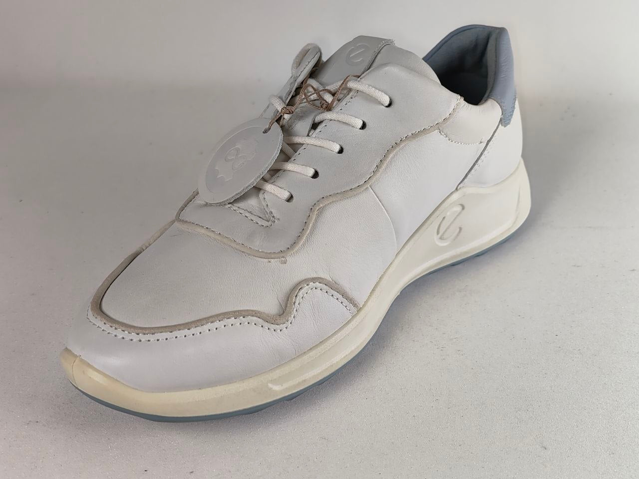 A white leather athletic sneaker with light gray accents, featuring a white laces and a thick white midsole with a light gray heel tab, photographed on a neutral light gray background.