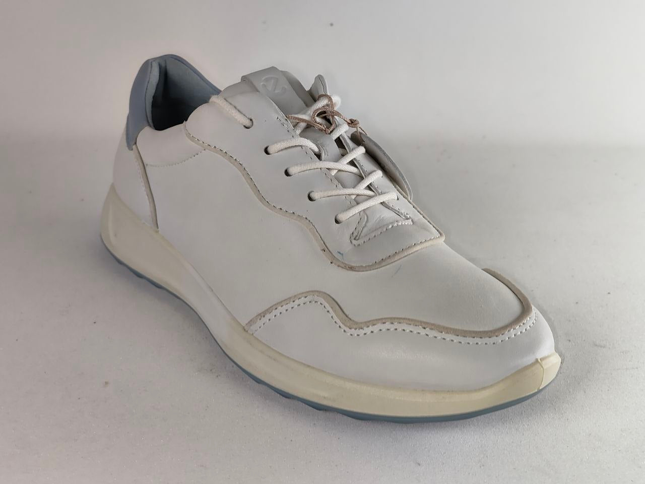 A white leather low-top sneaker with white laces, a light gray heel tab, and a cream-colored sole, shown on a neutral light gray background.