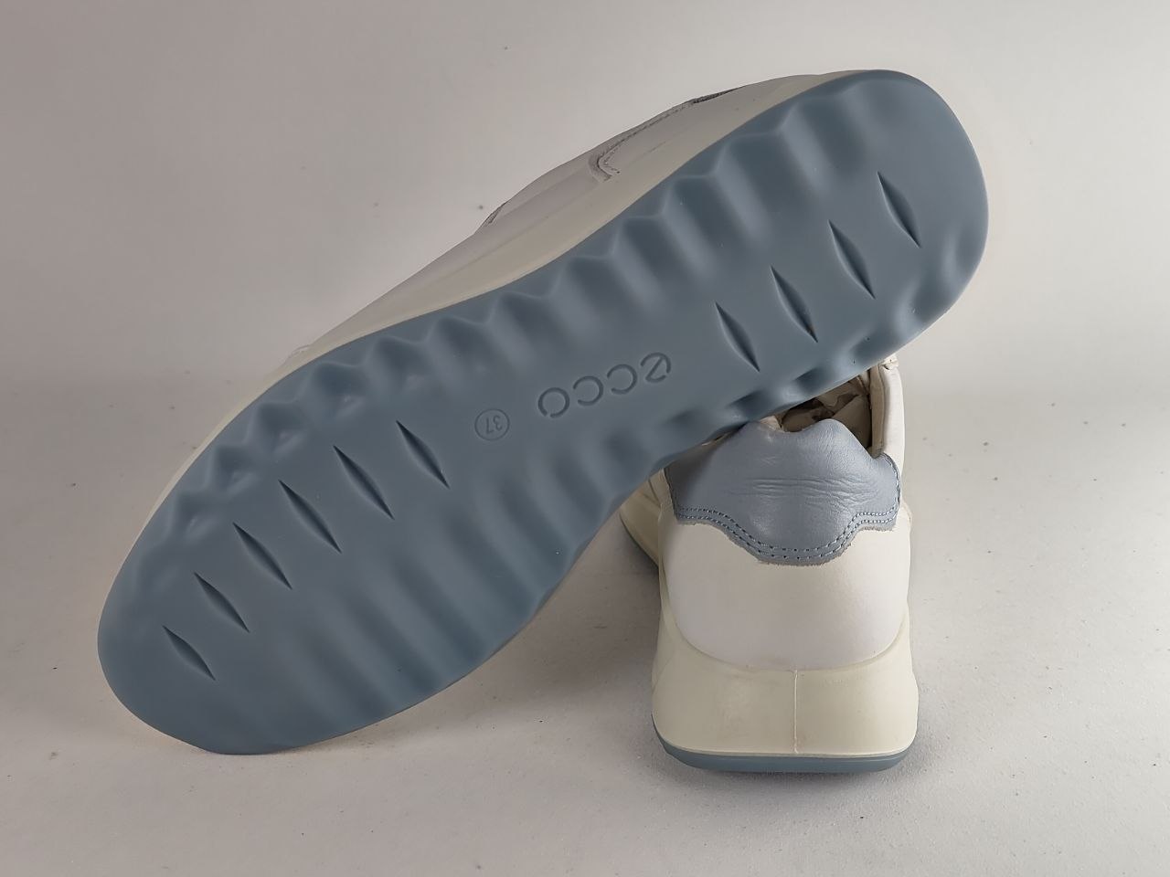 A white and light blue sneaker shown from the sole side, with the tread pattern and embossed 'ecco' logo visible, set against a plain light gray background.