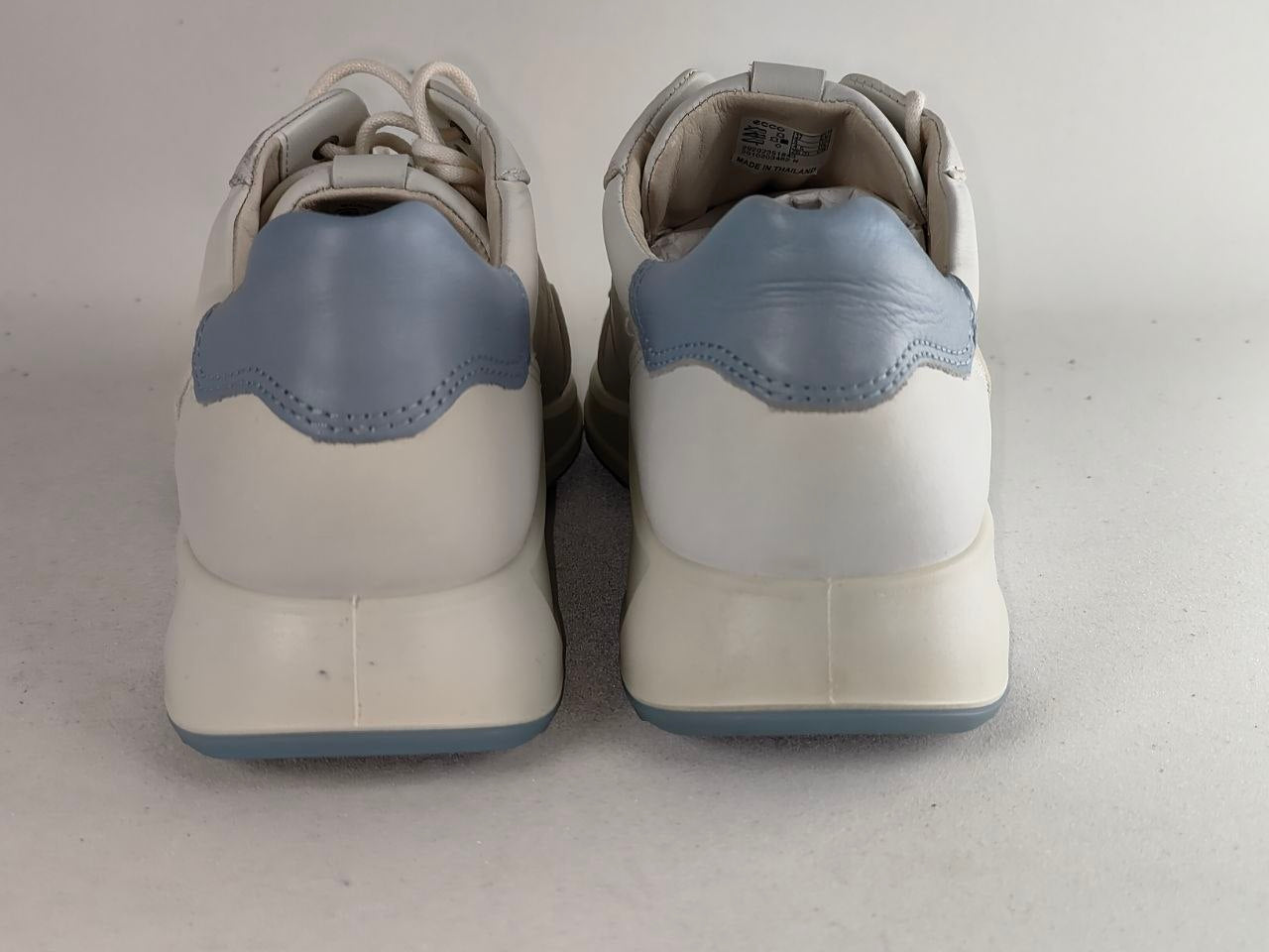 A pair of white low-top sneakers shown from the back, featuring light blue leather heel accents and matching light blue outsoles, set against a plain light gray background.