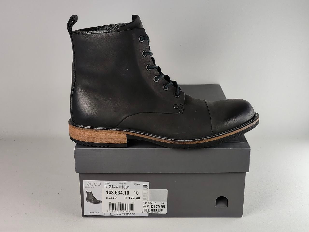 A black leather lace-up ankle boot with a tan sole, displayed on top of a gray ECCO shoebox against a light gray background.