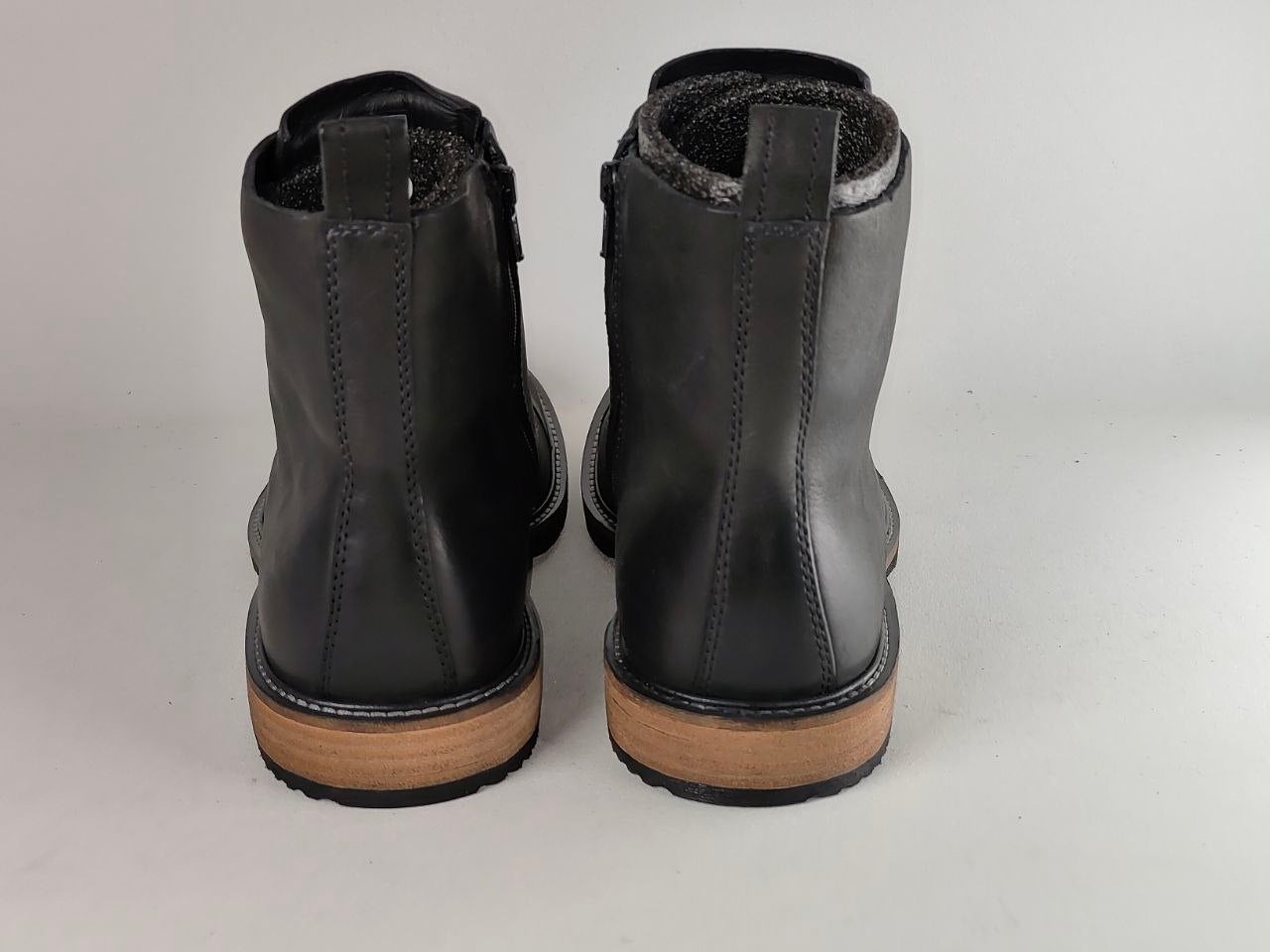 A pair of black leather ankle boots shown from the back, featuring a tan sole and a dark gray fuzzy lining visible at the collar, set against a plain light gray background.