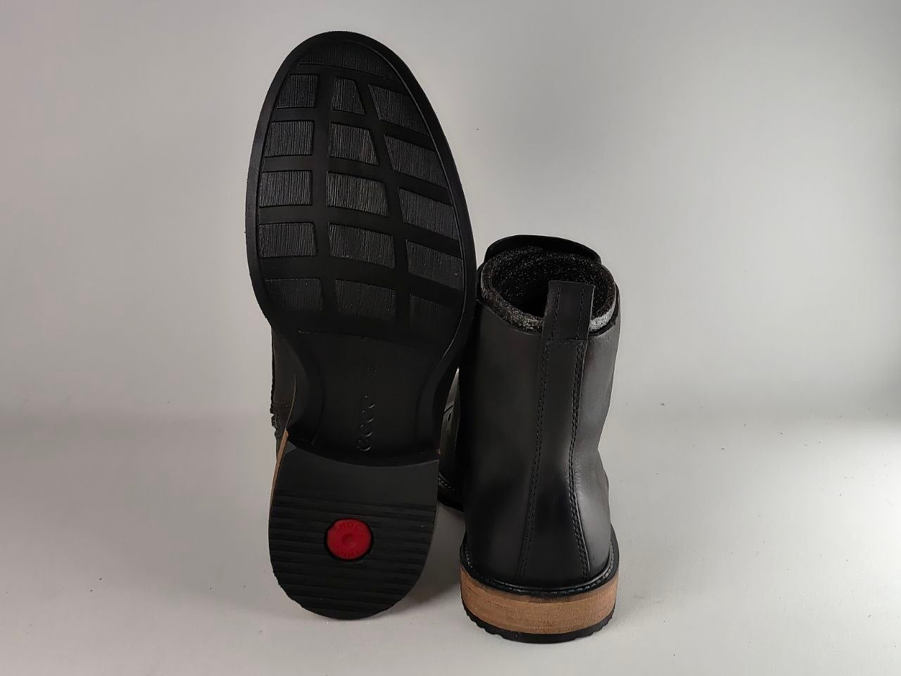 A pair of black leather ankle boots shown from the back, with one boot standing upright and the other tilted to display the sole. The sole features a black rubber tread with a small red circular accent near the heel, and a light tan outsole.