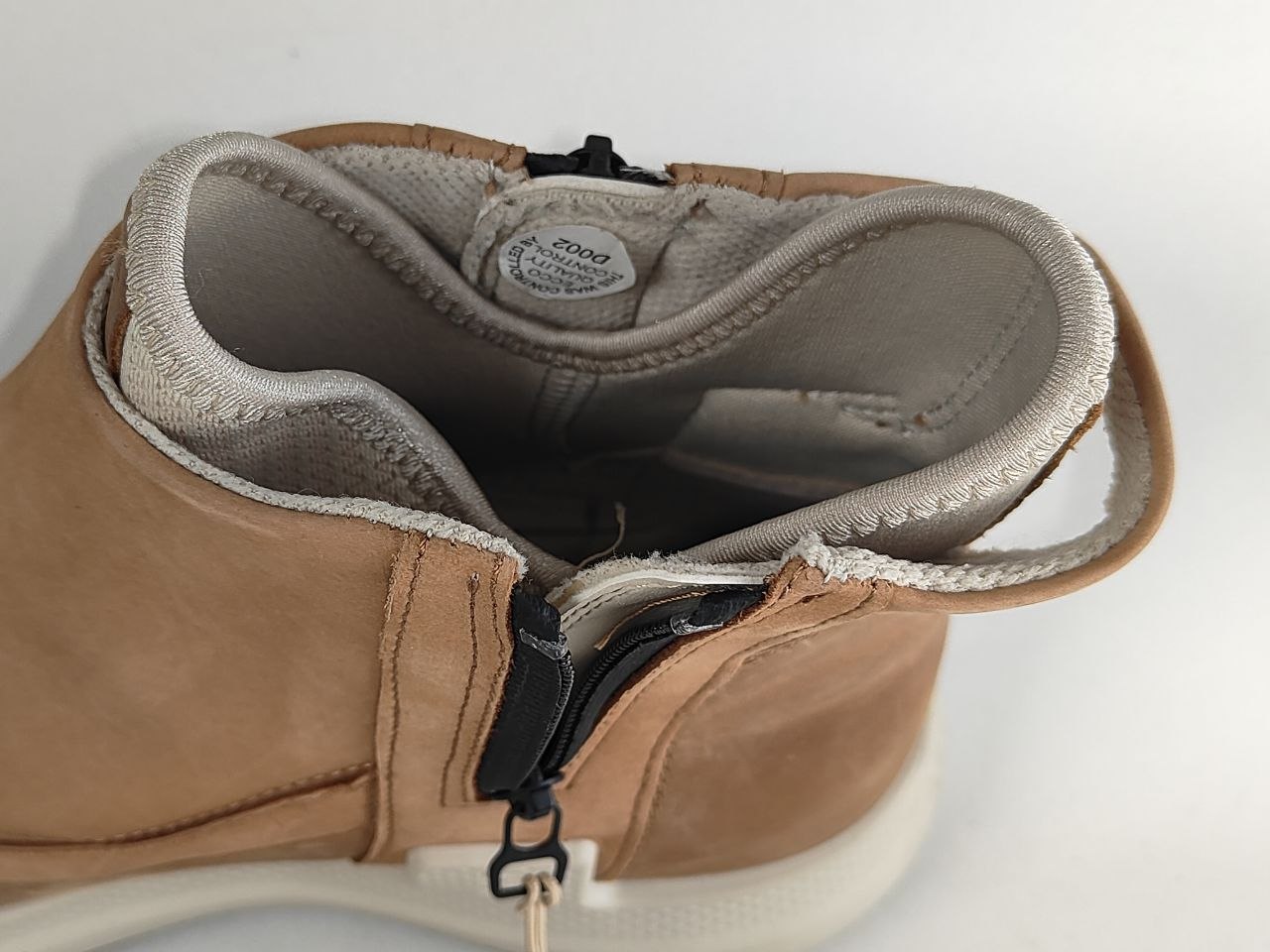 A close-up of a tan suede ankle boot with a side zipper, showing the interior lining and a white sole against a white background.