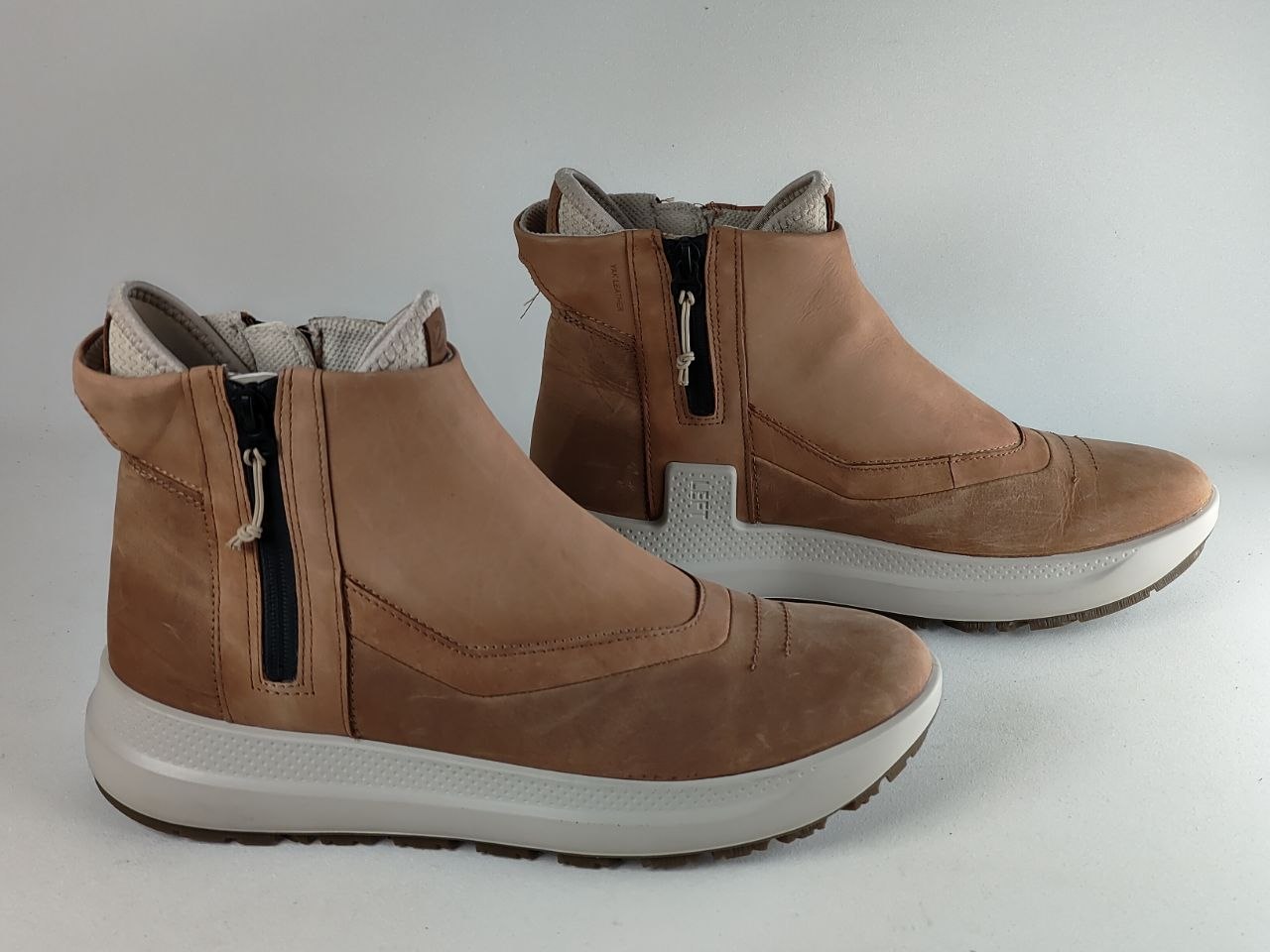 A pair of tan suede and faux-suede ankle boots with side zippers, white midsoles, and dark outsoles, shown against a plain light gray background.