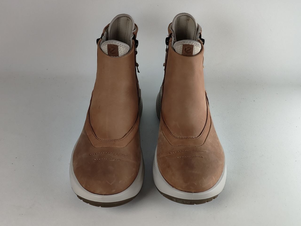 A pair of tan leather ankle boots with white soles, photographed from the front against a neutral light gray background.