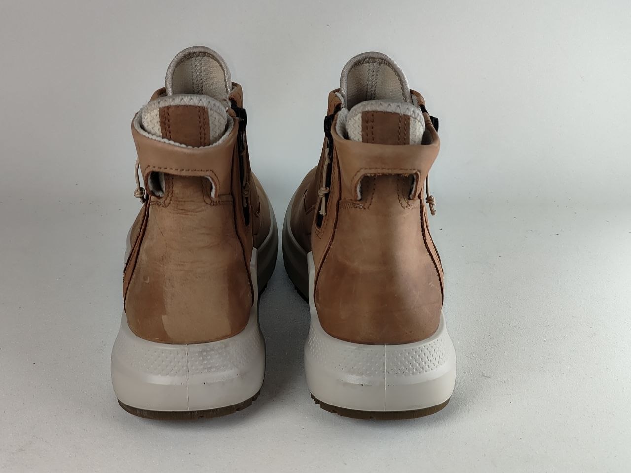 A pair of tan leather high-top boots with white soles, shown from the back against a light gray background.