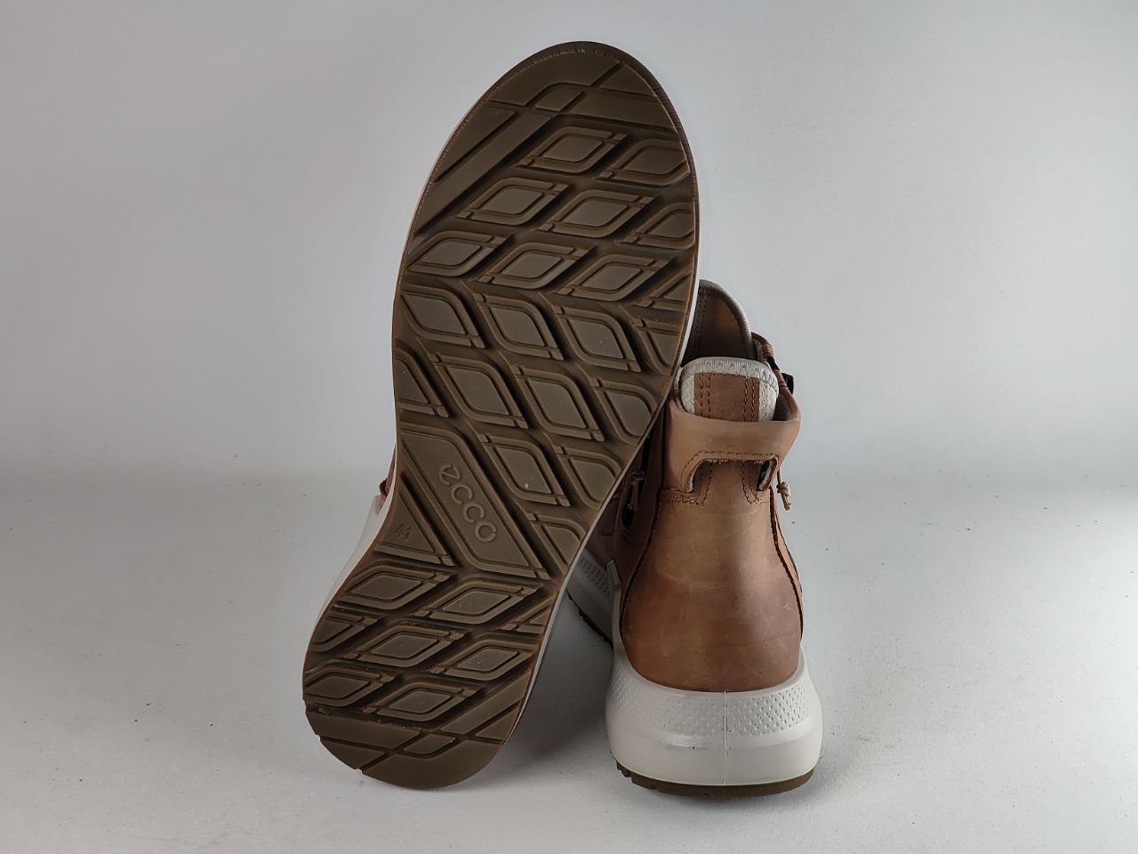 A pair of brown leather ankle boots shown from the side and sole, featuring a white midsole and a textured brown outsole with the brand name 'ecco' visible.