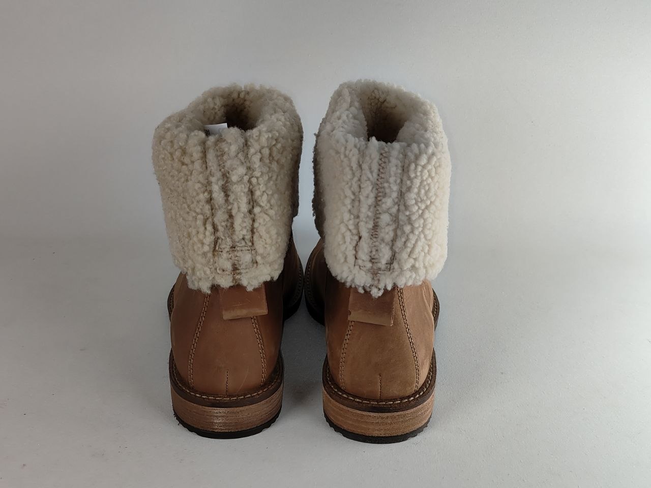 A pair of mid-calf brown boots with thick cream-colored shearling or fleece cuffs, shown from the back against a plain light gray background.