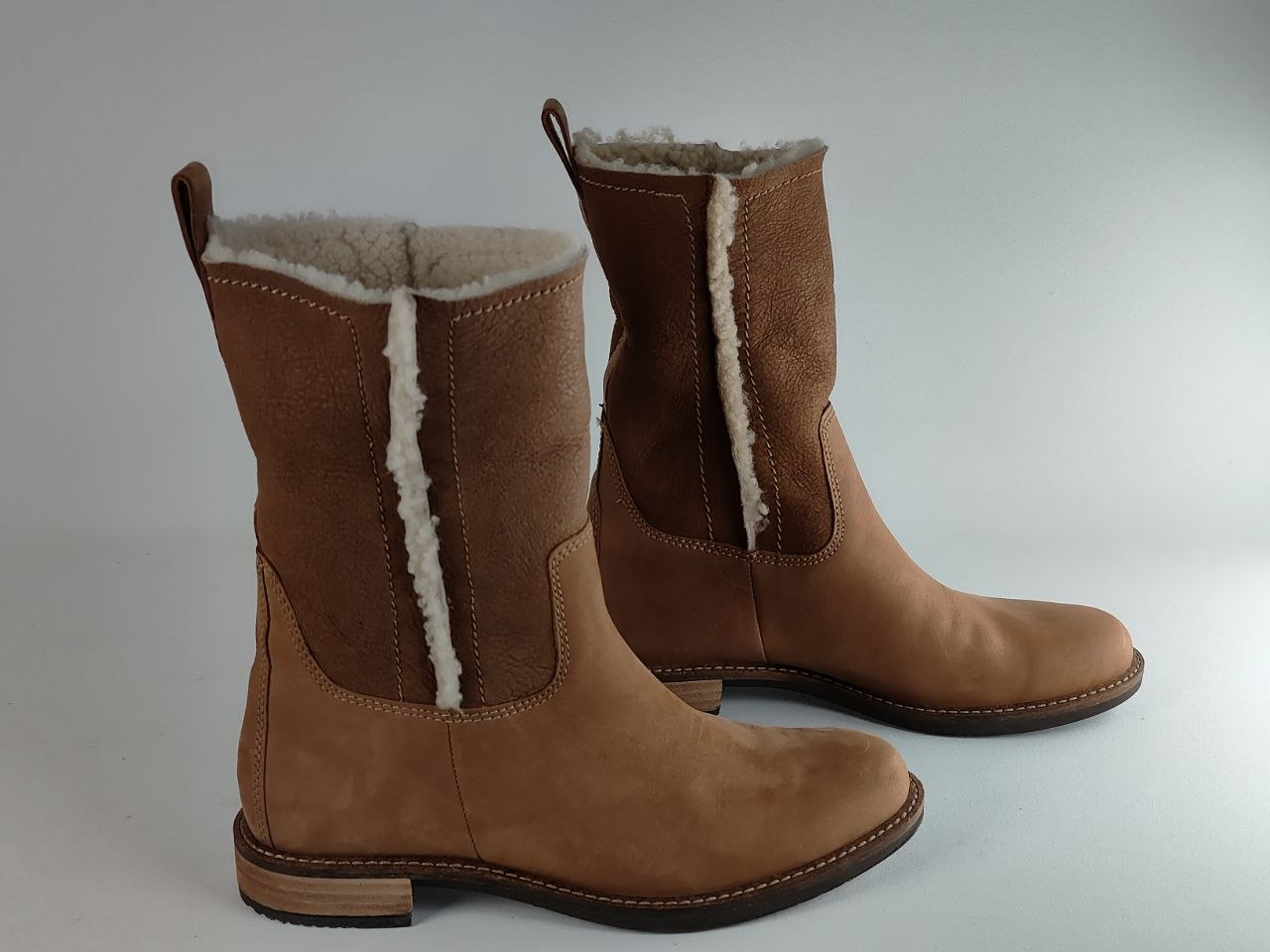 A pair of mid-calf brown suede boots with white shearling lining, displayed on a light gray background.