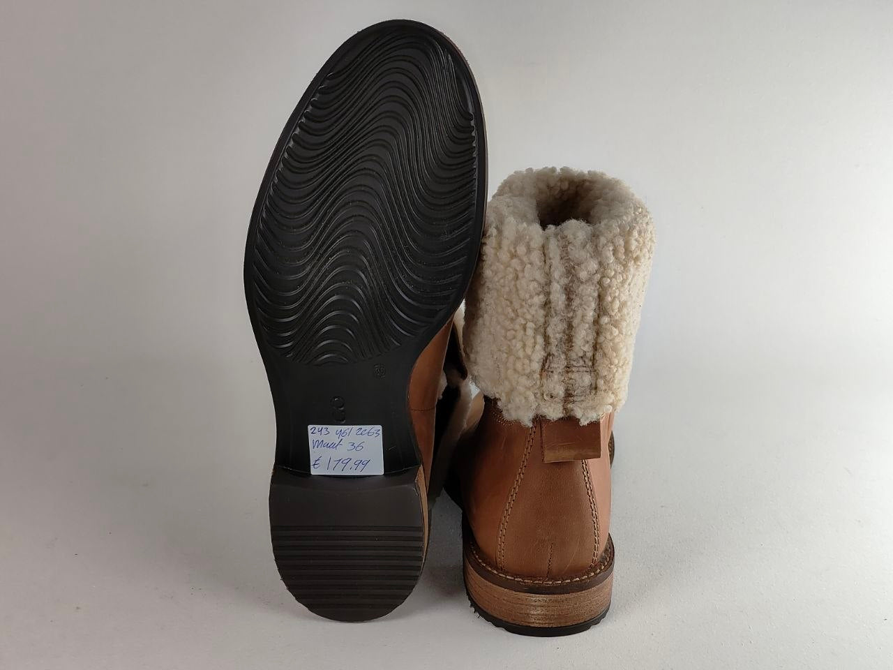 A pair of brown leather ankle boots with a thick cream-colored shearling or fleece lining, shown from the side and sole. A small white size tag is visible on the black outsole.