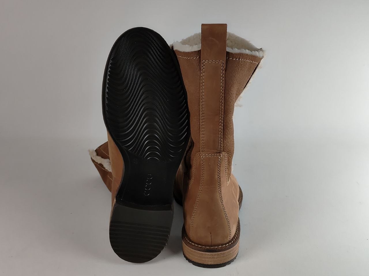 A pair of tan suede boots with white shearling lining, shown from the back to display the sole tread and the inner lining.