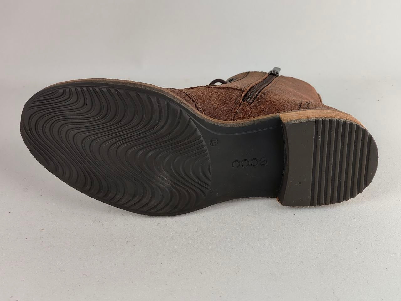 A brown suede ankle boot shown from the sole view, featuring a black treaded rubber outsole and a small metal zipper near the top.