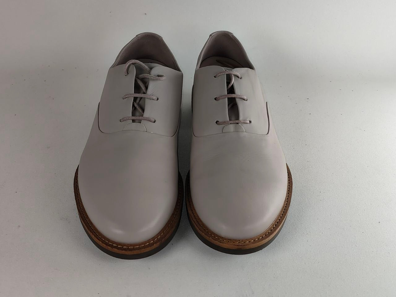 A pair of light gray leather lace-up shoes with brown soles, photographed from the front on a plain light gray background.